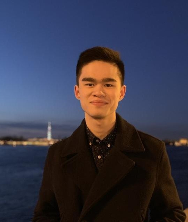 A young man with black hair and a black coat stands in front of a body of water at night