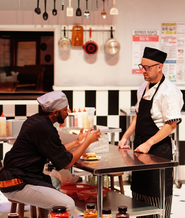 on a stage in a restaurant kitchen, a Black man sits at a table while talking to a white man standing