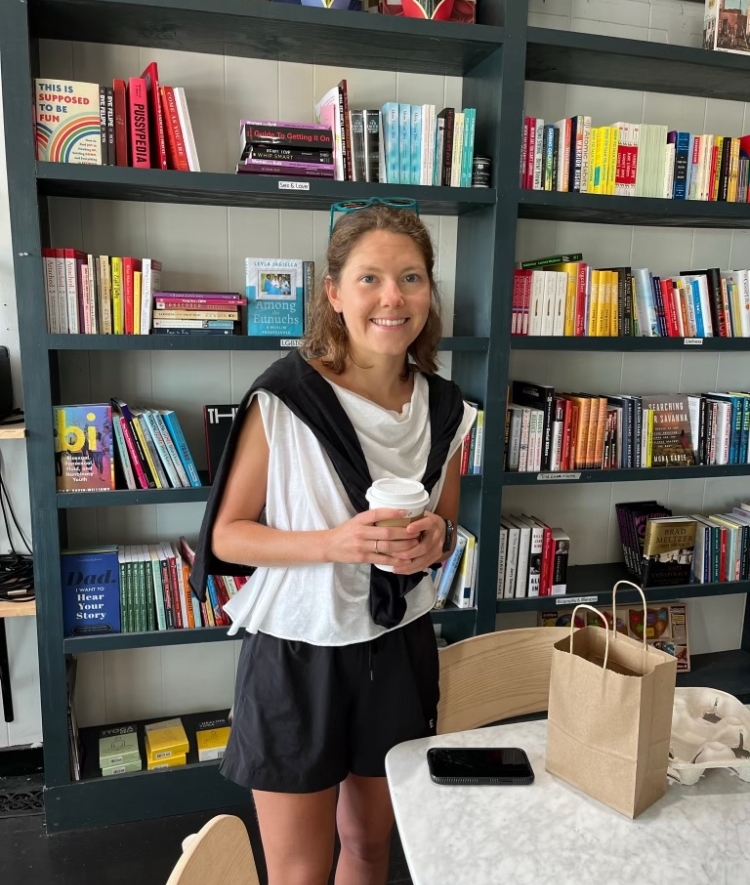 a young woman wearing a white tank and black shorts holds a coffee while standing in a bookstore