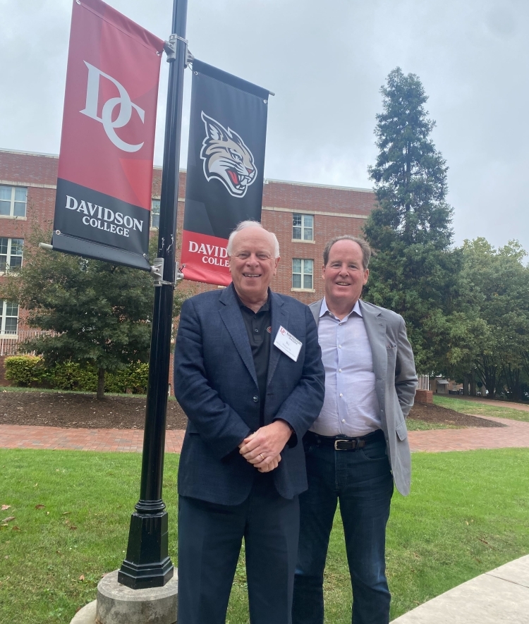 two men standing in front of a grassy lawn with a lamp in the background holding a "Davidson College" sign