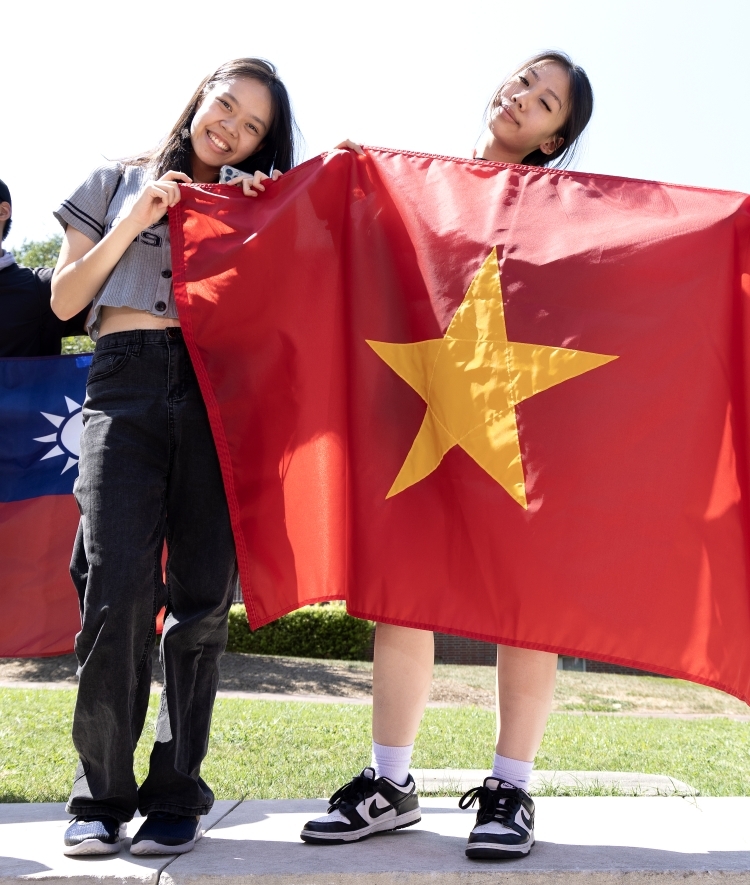 Two young women hold flags of various countries while smiling