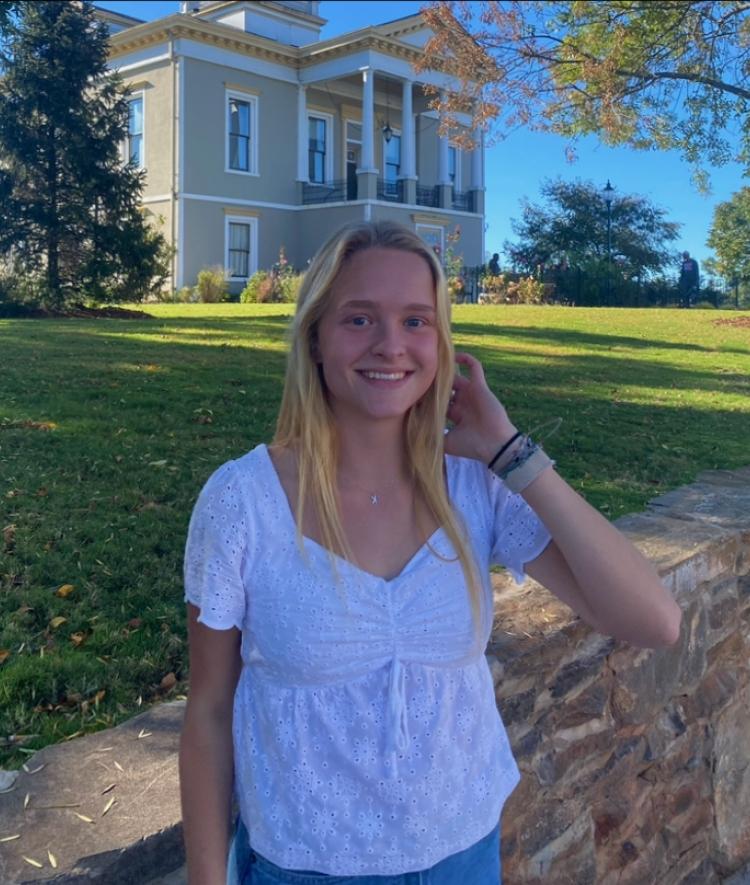a young woman with blonde hair wearing a white top stands in front of a historic house on a sunny day