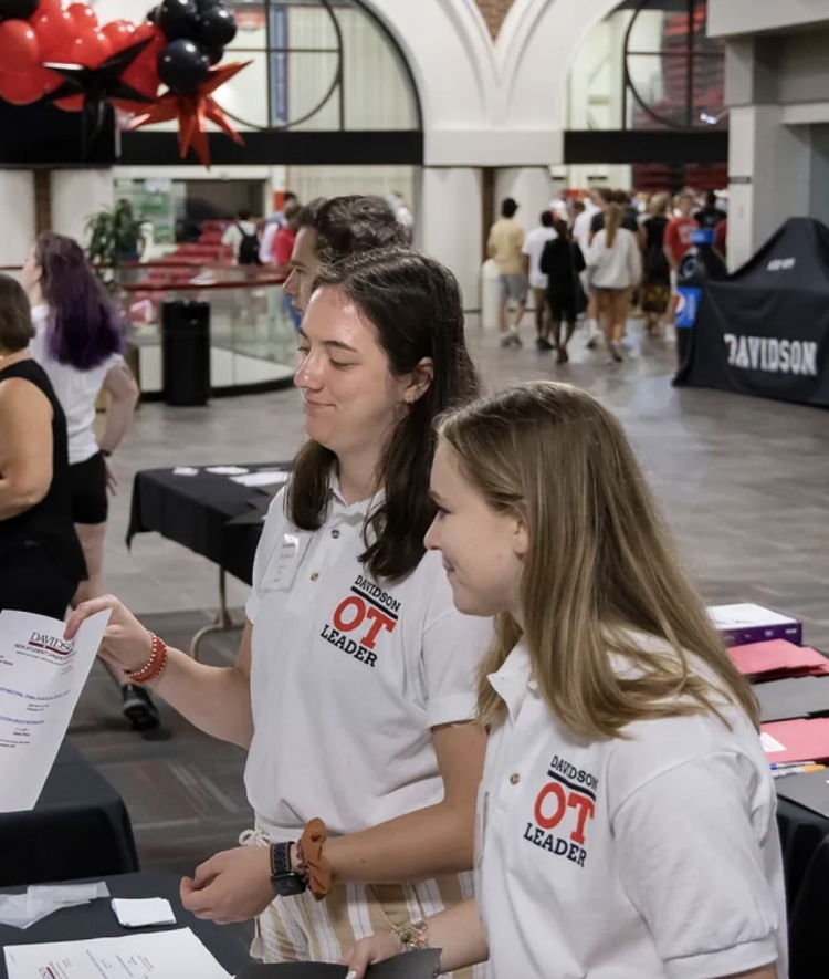 two young women in white polos talk to an older woman around a table with balloons in the background