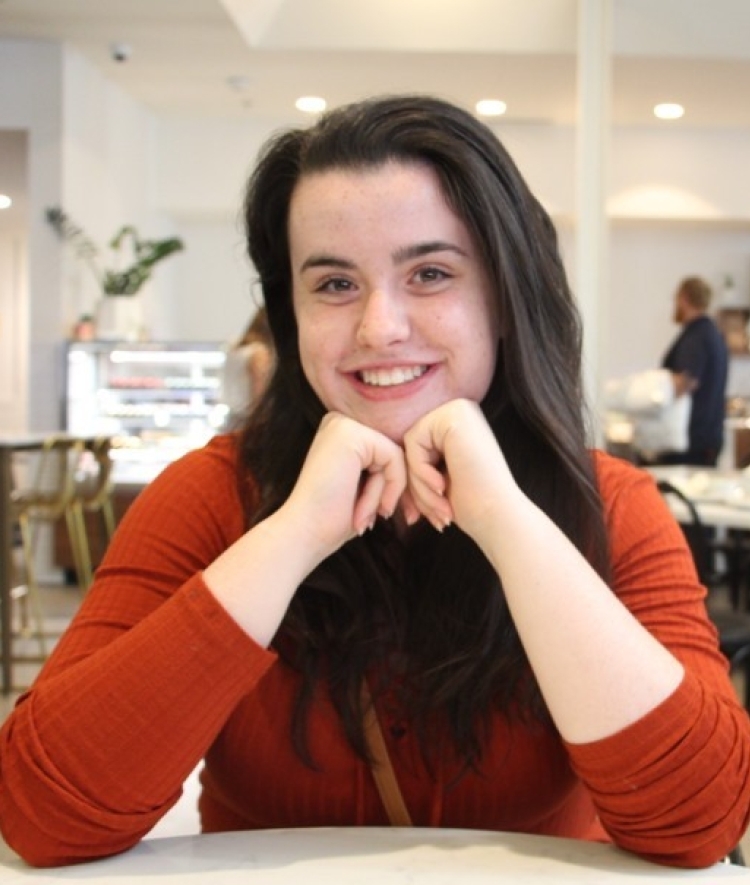 a young woman with brown hair wears an orange shirt and smiles