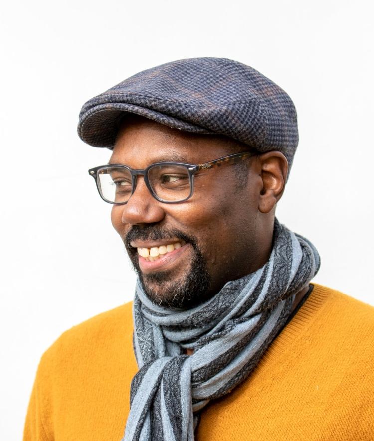 a young Black man wearing a yellow sweater and a striped scarf with a hat while smiling