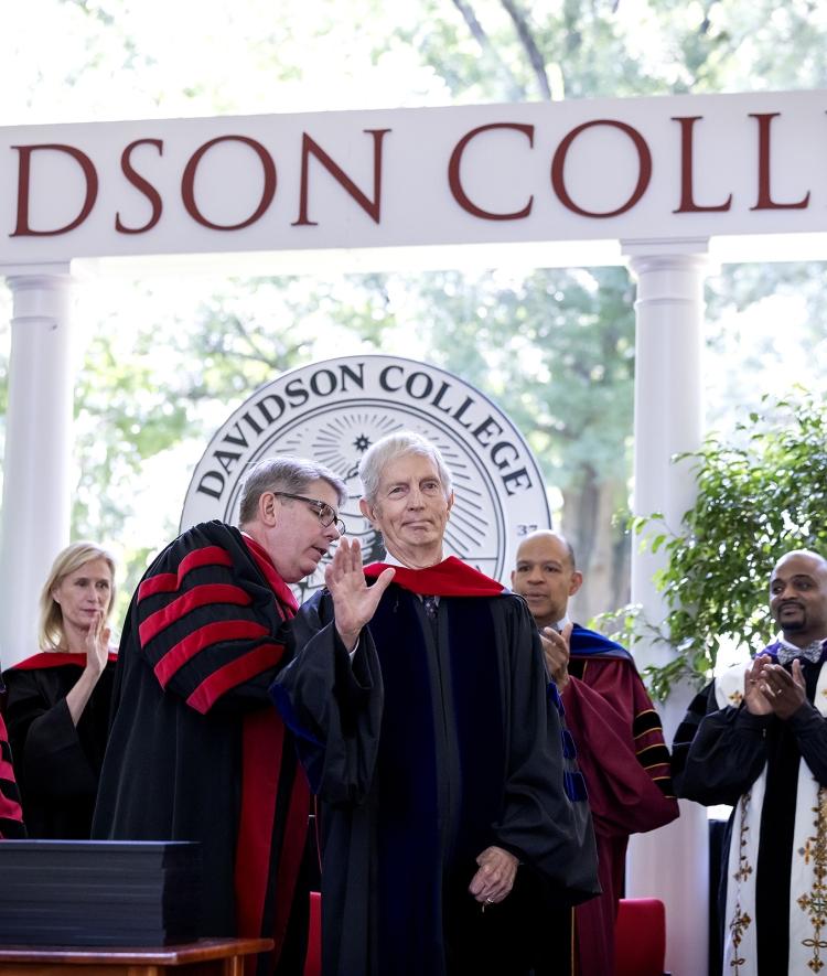 an older white man puts academic regalia on an older white man while on a stage