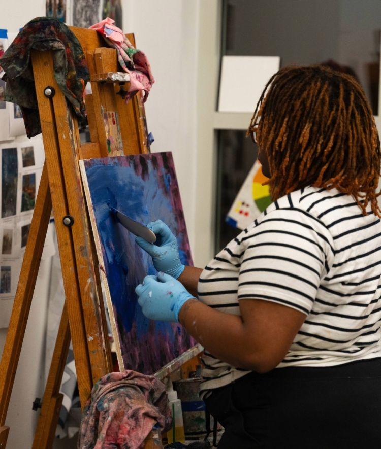 a young Black woman paints at an easel in an art studio