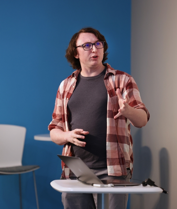 a young white man wearing a flannel and speaking in front of a crowd