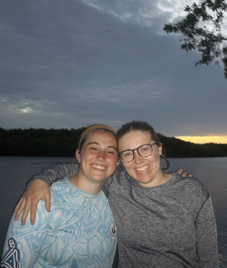 two young women standing in front of a body of water at sunset