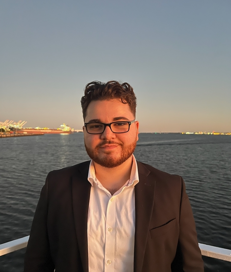 a young Middle Eastern man wearing glasses and a jacket in front of a body of water at sunset