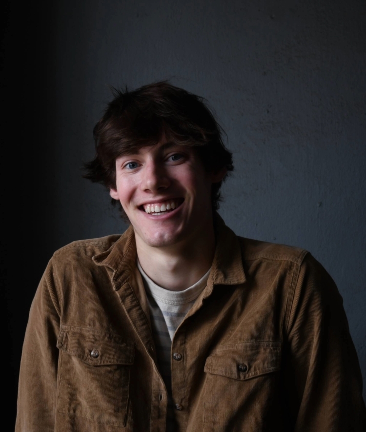 a young white man with dark hair wearing a brown khaki shirt and smiling on a grey background