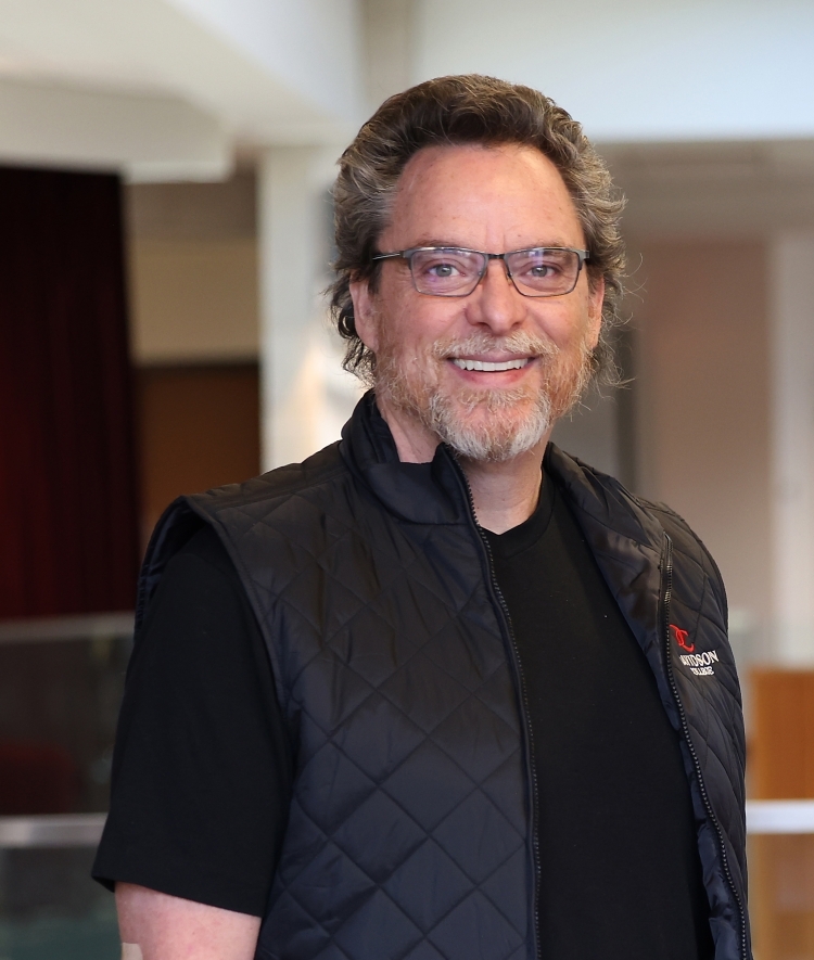 an older white man wearing a black shirt and a black vest with glasses