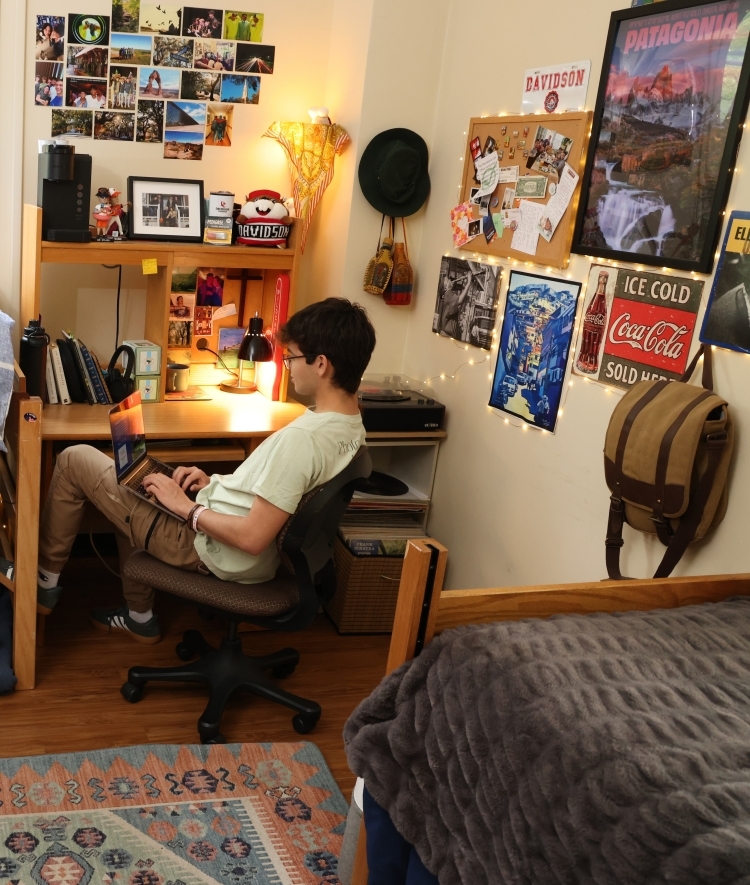 a young man works at his computer at a desk in a dorm room