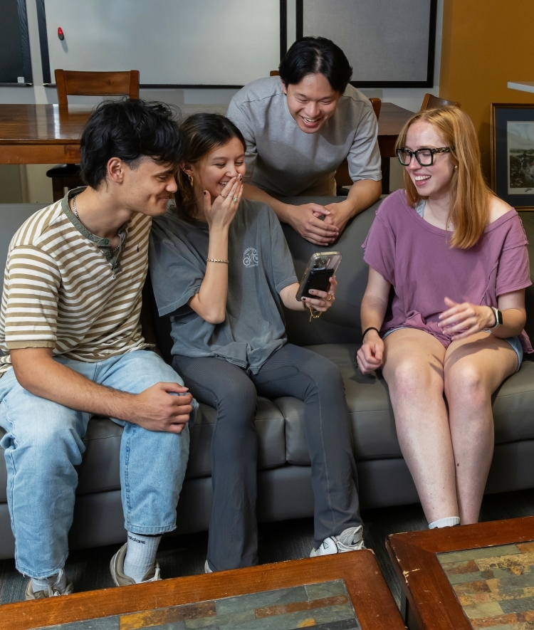 a group of four students sit together on a couch in a lounge laughing at a phone
