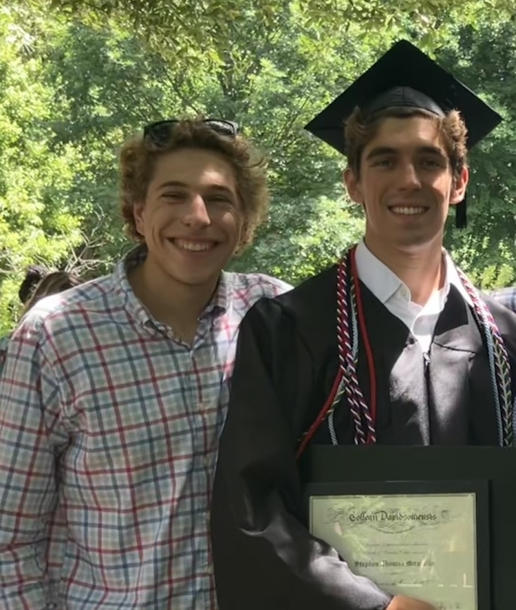 two young men stand together as one graduates in regalia holding a diploma on a sunny day
