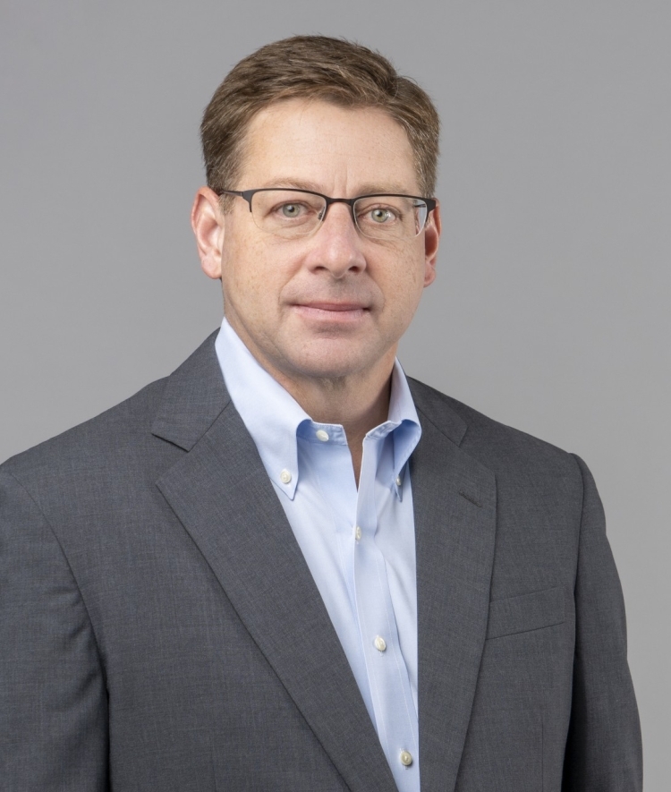 a middle aged white man with dark hair wearing glasses, a blue collared shirt and a grey jacket