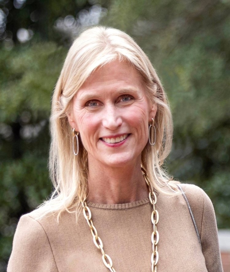 a white woman with blonde hair wearing a tan top and gold necklace smiling outside