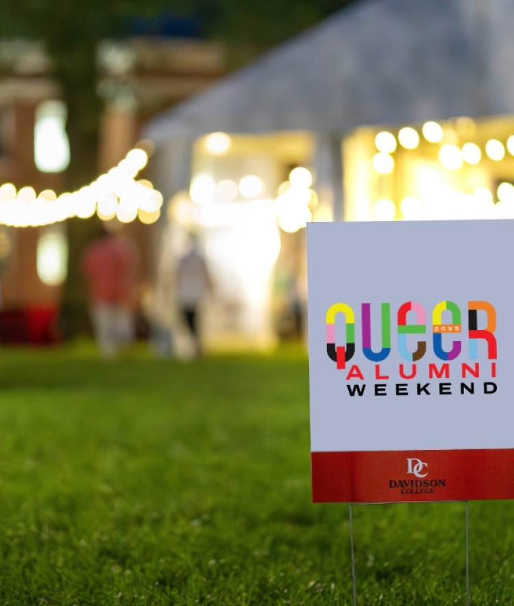 a lawn sign that reads "Queer Alumni Weekend" in front of a large white tent with string lights at night