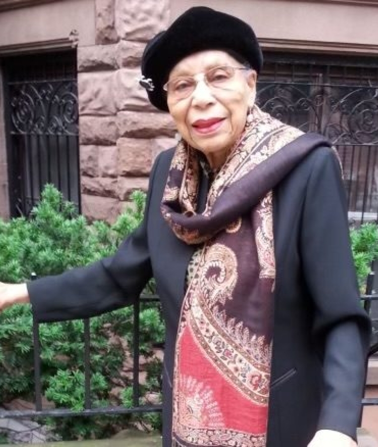 a Black woman wearing a cap and scarf standing in front of a brownstone