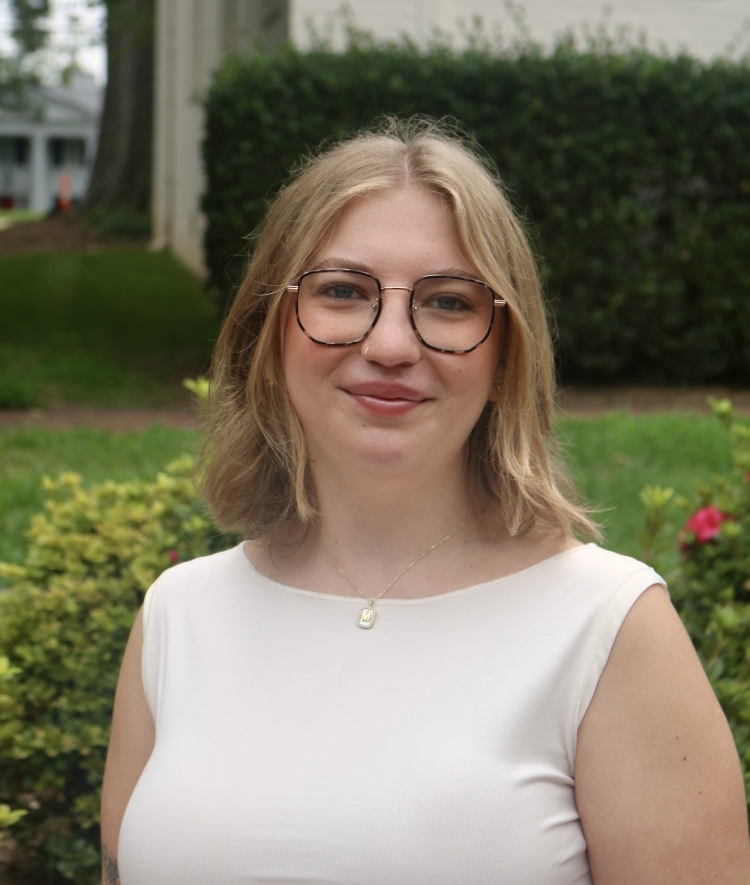 a young woman with short blonde hair wearing glasses and a white top smiling outside