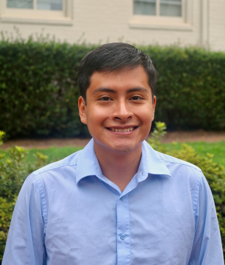 a young man with short dark hair wearing a blue collared dress shirt and smiling outside