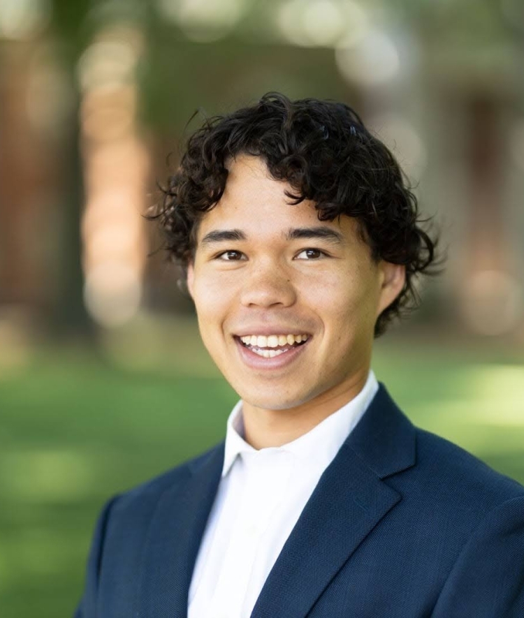 a young man with dark curly hair wearing a collared white shirt and navy blazer