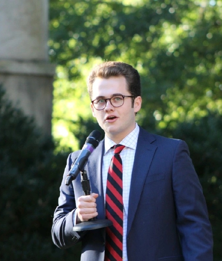 a young man with short brown hair wearing a suit and tie with glasses while holding a microphone outside