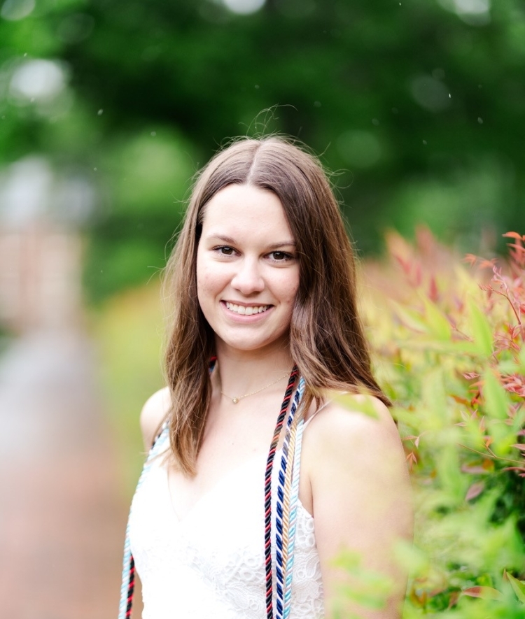 a young woman with brown hair wearing a white dress and graduation cords smiling outside