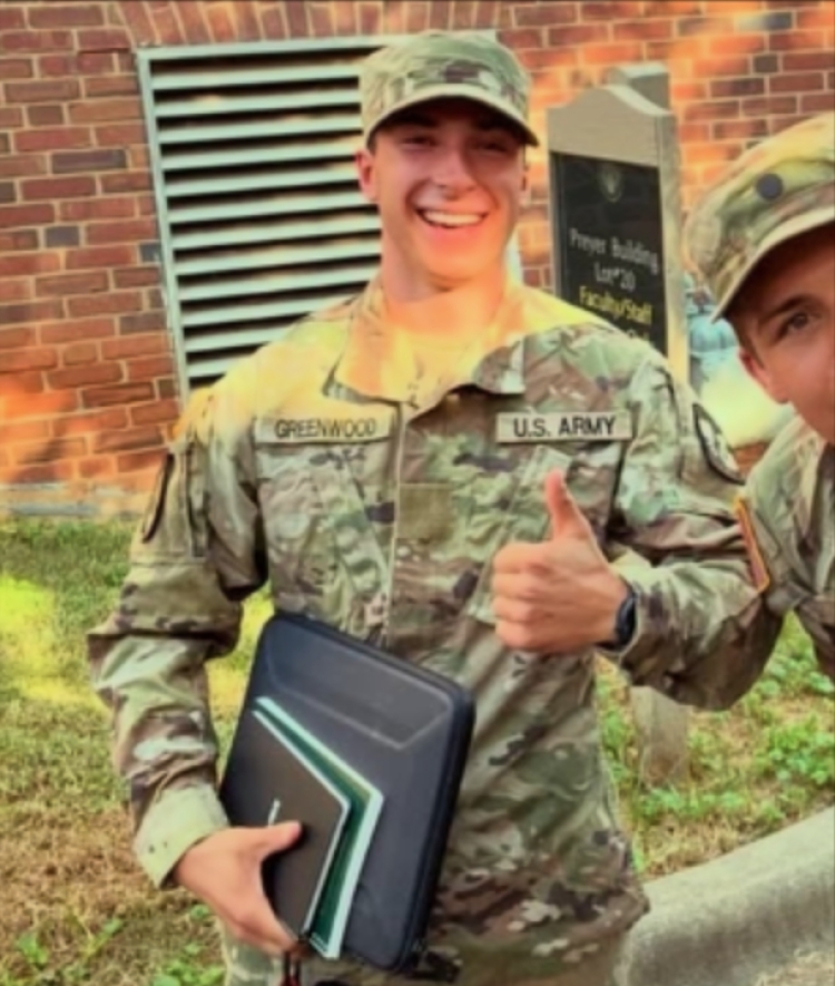 A smiling college age man in military fatigues holding a stack of books and giving a thumbs up
