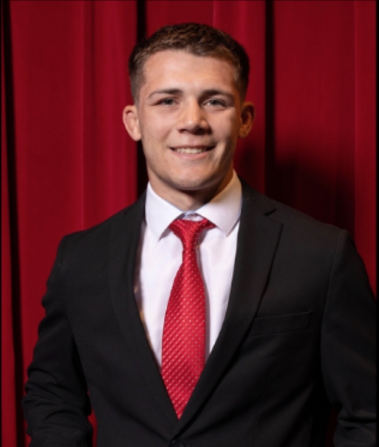 College-age male wearing a black blazer and red tie in front of a red curtain