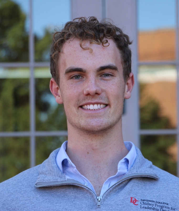 a young white man with curly hair wearing a half zip sweatshirt and collared shirt smiling