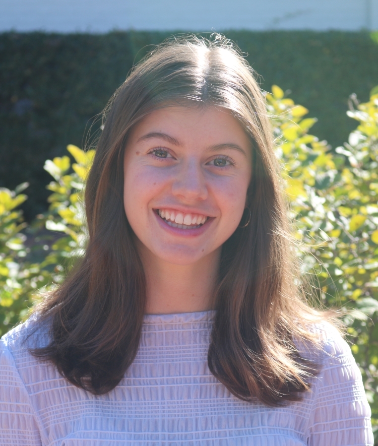a young woman with brown hair wearing a white top smiling outside on a sunny day