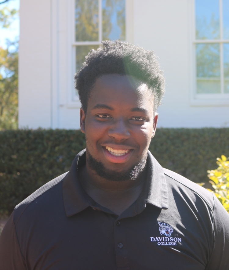 a young Black man wearing a black collared shirt smiling outside on a sunny day