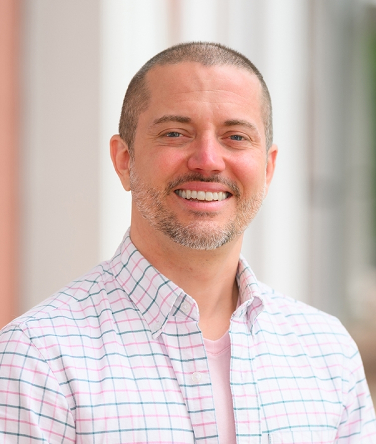 An adult man smiles with a shaved head and dark greying facial hair