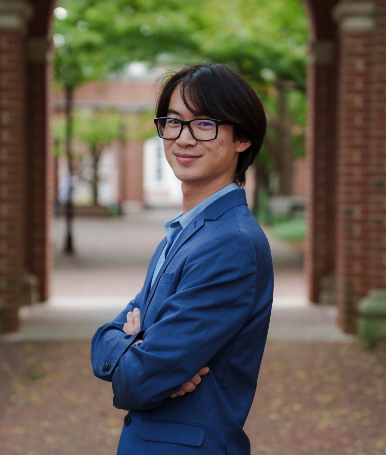 A young man with glasses and dark hair smiles while standing with crossed arms in a blue blazer, positioned outdoors between two large brick pillars on a college campus.