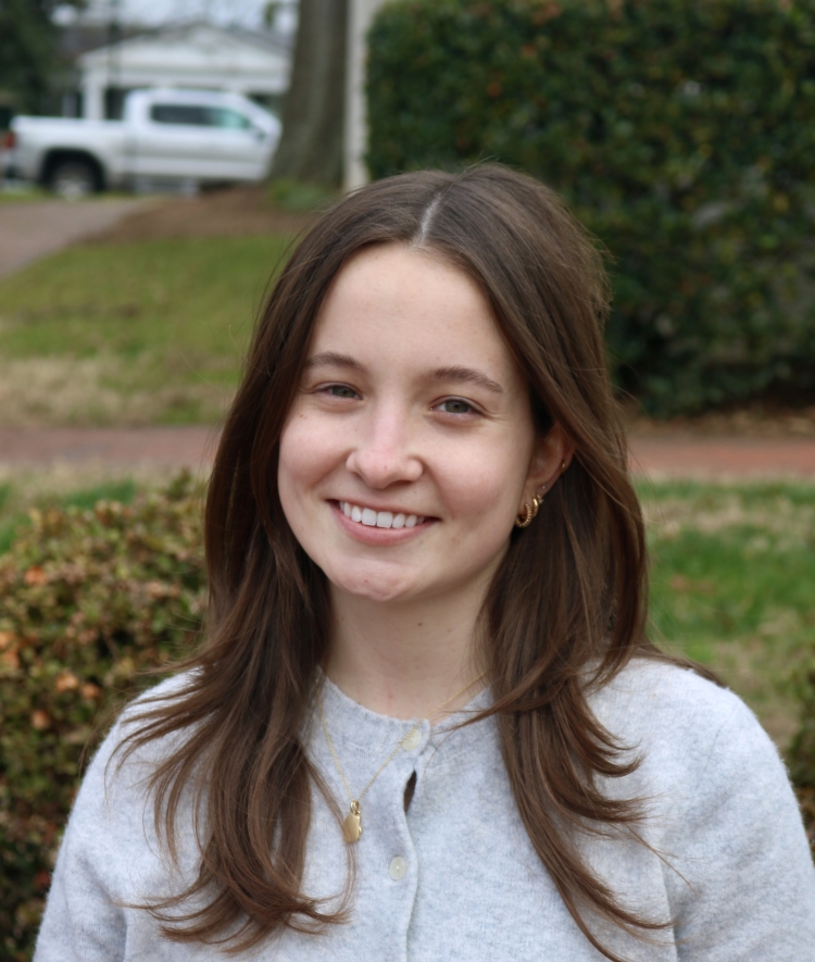 A young woman with long brown hair smiling outdoors while wearing a light gray button-down cardigan and a gold necklace.