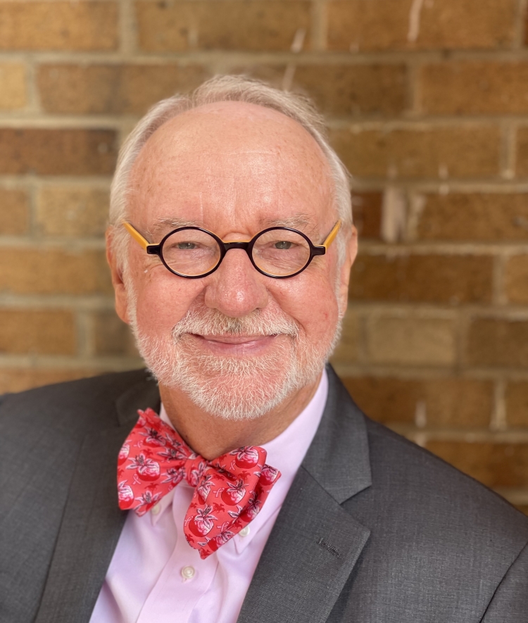 Don Davis wears a grey suit, pink shirt, and a vibrant red floral bowtie while smiling in front of a brick wall.