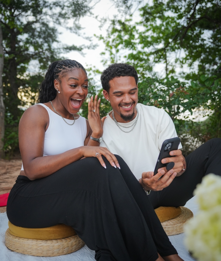 A young woman reacts with a joyful, open-mouthed expression while showing off an engagement ring as she and a smiling young man look at a smartphone together outdoors.