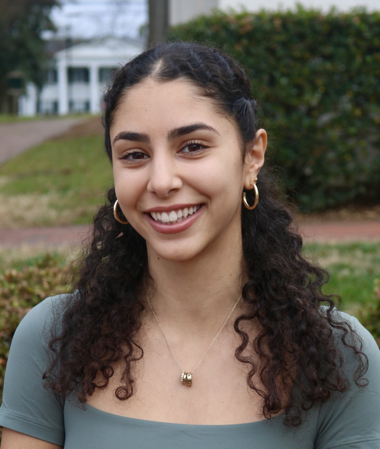 A young woman with curly dark hair and gold hoop earrings smiling outdoors while wearing a sage green fitted top and a gold pendant necklace.