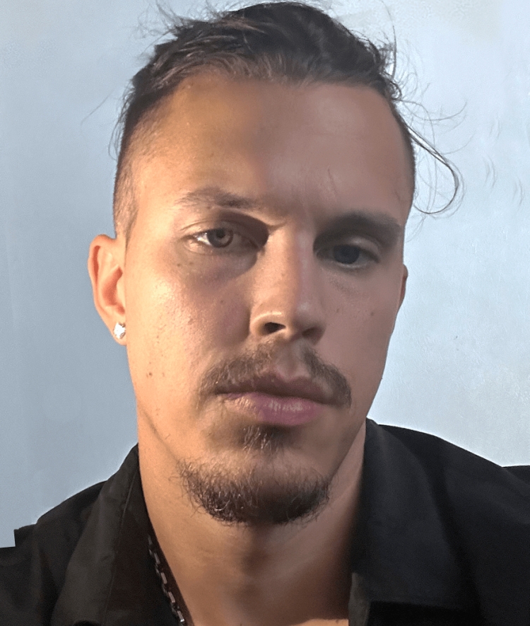 A man with a goatee, mustache, and dark hair pulled back looks directly into the camera while wearing a black collared shirt and a silver chain against a light-colored background.