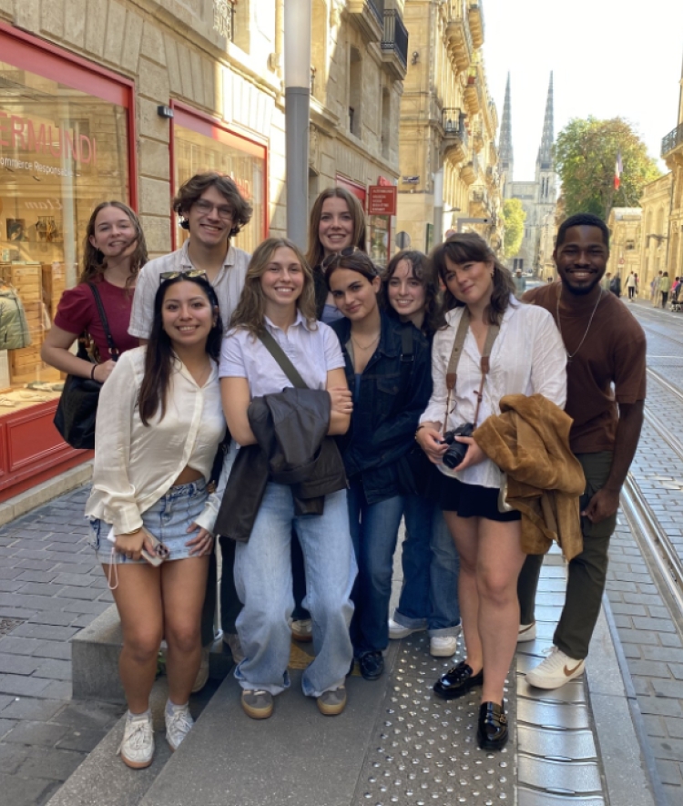A diverse group of nine smiling students poses on a cobblestone street in Bordeaux next to an "Altermundi" storefront, with tram tracks and distant cathedral spires visible in the background.