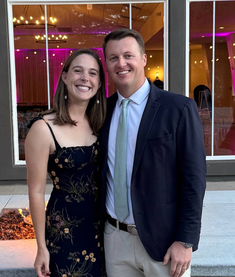 A smiling couple stands together on an outdoor patio in front of a stone fire pit and a building with large windows reflecting purple interior lighting.
