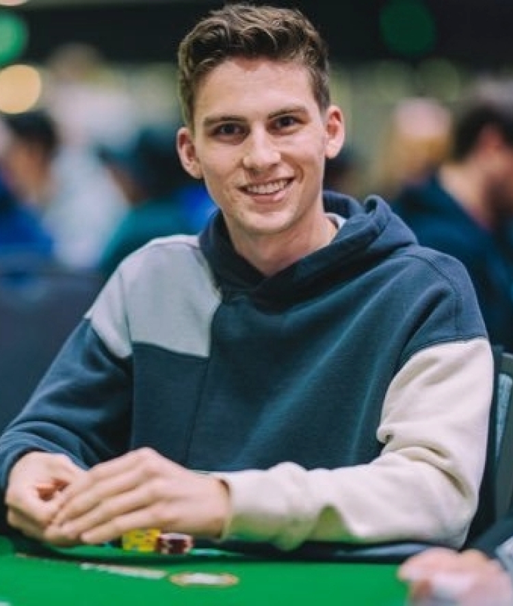 A smiling young man with brown hair wearing a color-blocked blue and grey hoodie sits at a green poker table with chips.