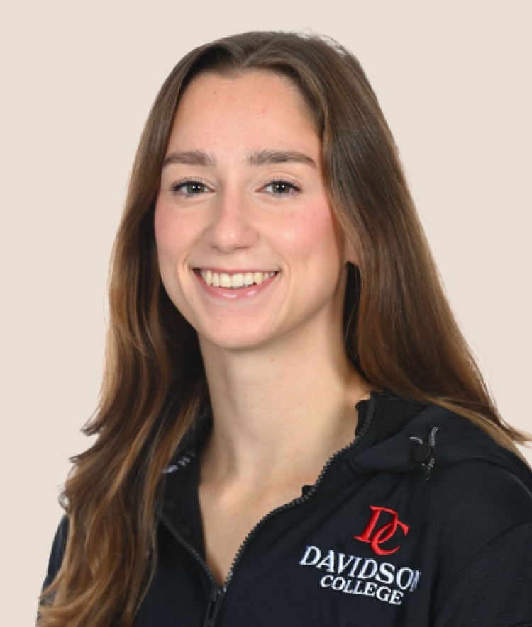 A smiling woman with long brown hair wears a black zip-up jacket featuring the Davidson College logo.