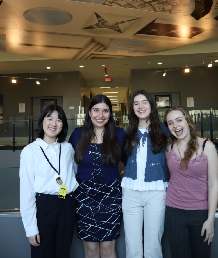 Four young women stand in a row smiling for a group photo under a decorative ceiling featuring scientific illustrations and symbols.