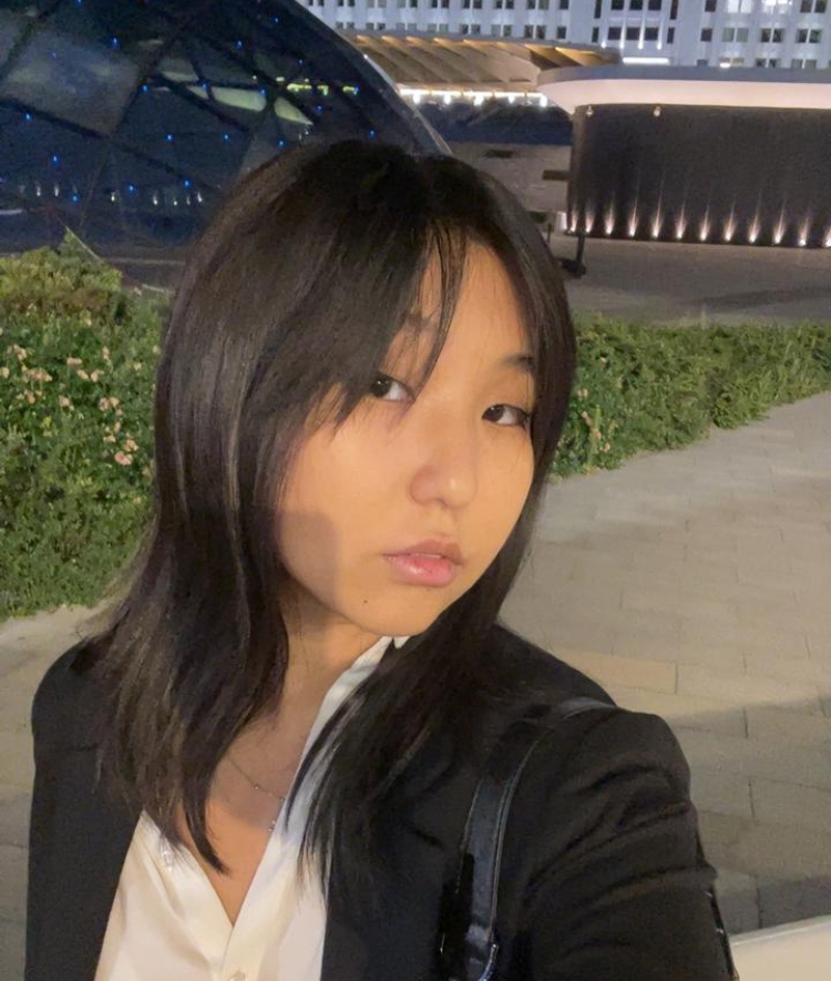 young woman with shoulder-length dark hair and a black blazer poses for a selfie at night in front of a modern building with blue lighting.