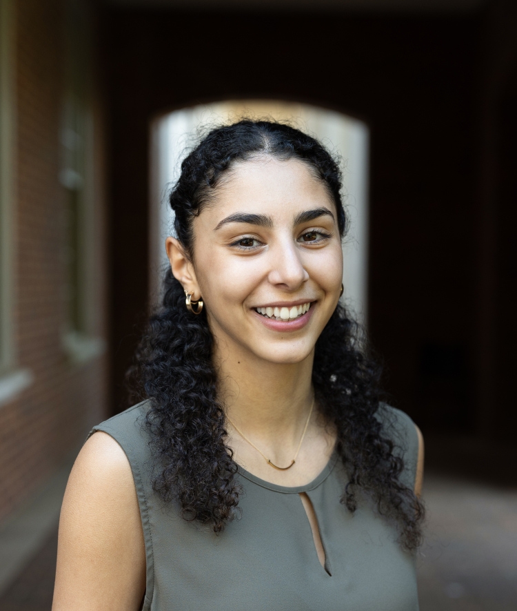 Maya Rajeh smiles in a professional headshot while wearing an olive green sleeveless top and gold jewelry.