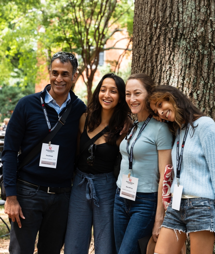 A smiling family of four poses together under a large tree during a Davidson College orientation event, with a background of outdoor tables and green foliage.