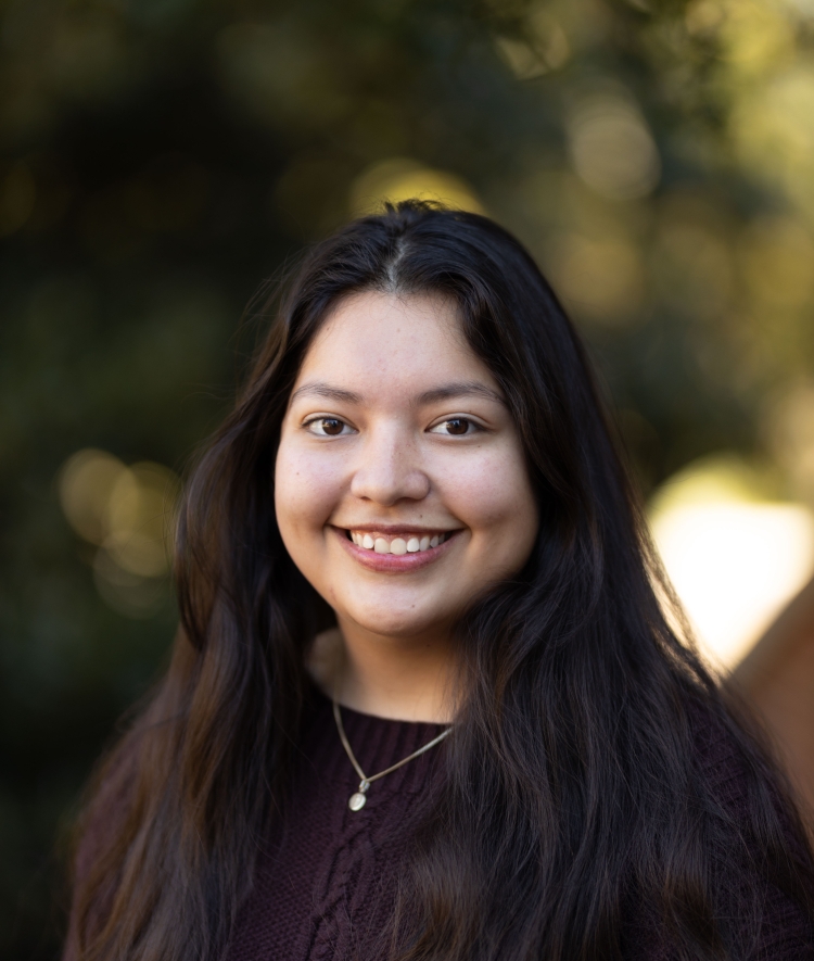 A smiling young woman with long, dark hair and a gold necklace poses for a headshot against a blurred outdoor background of green foliage.