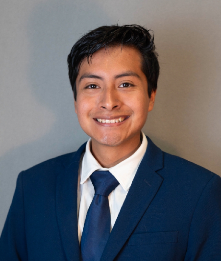 A professional headshot of a smiling young man with dark hair wearing a navy blue suit, white shirt, and blue tie against a grey background.